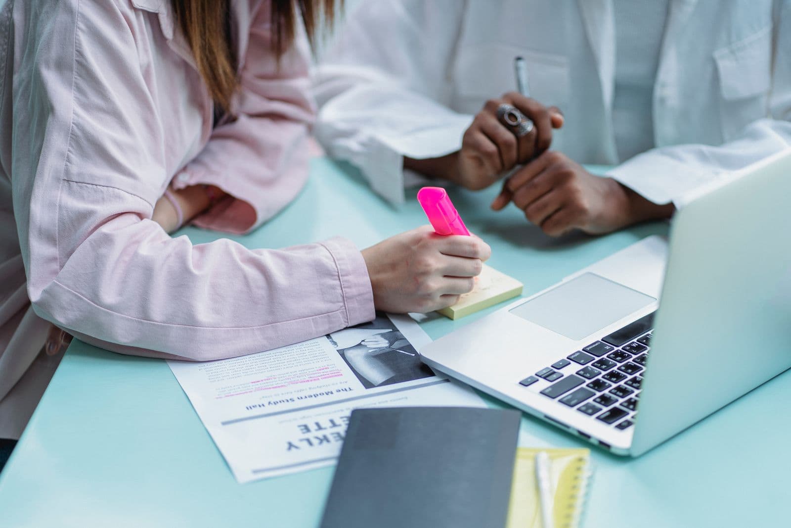 Two researchers at a desk, one highlighting findings in a printed article and writing codes on a sticky note beside a laptop